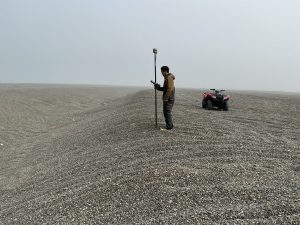 Someone stands on a small hill in a field of gravel. They are holding a Trimble instrument. There is an ATV nearby. The sky is misty and grey.