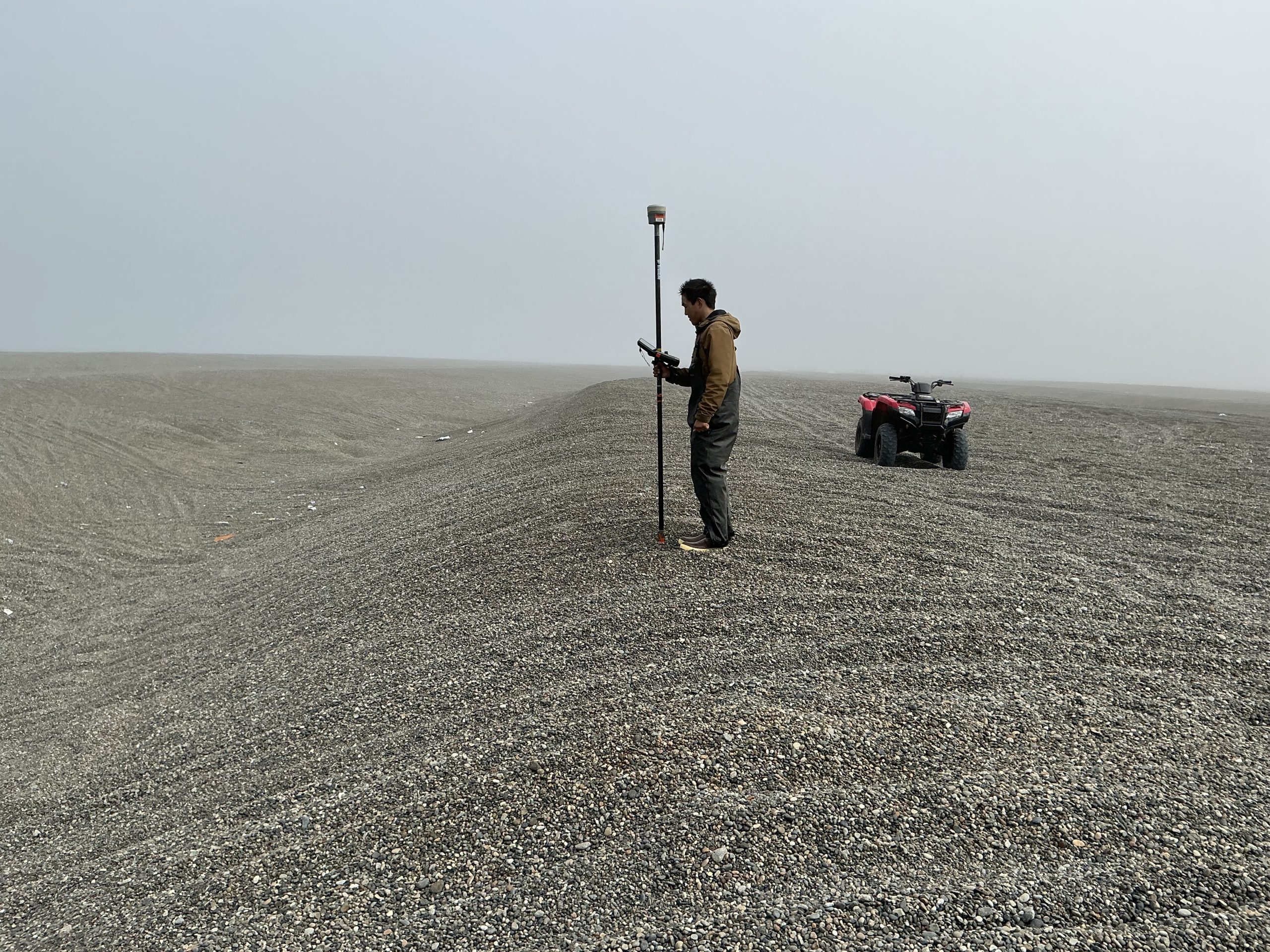 Someone stands on a small hill in a field of gravel. They are holding a Trimble instrument. There is an ATV nearby. The sky is misty and grey.