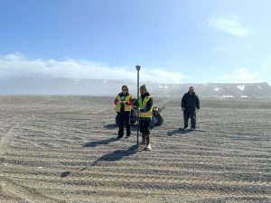 Three people stand nearby each other. One is holding a Trimble instrument, another is holding a notebook, and the thrid is walking towards the other two. There is an ATV nearby them. They are standing in a gravelly field covered in tire tracks. There is a cliff behind them that has patches of snow and obscured by mist. The sky is partially cloudy.