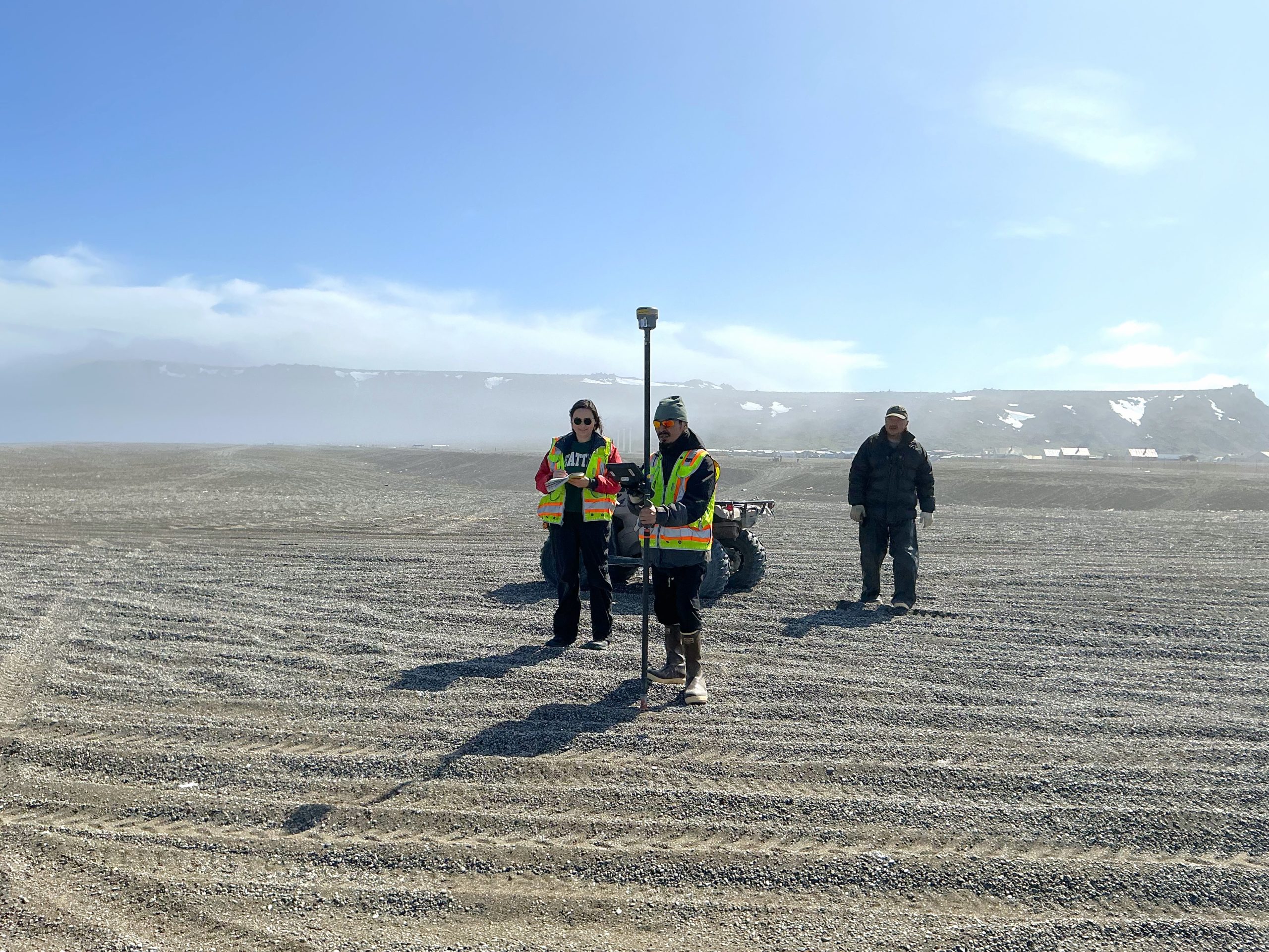 Three people stand nearby each other. One is holding a Trimble instrument, another is holding a notebook, and the thrid is walking towards the other two. There is an ATV nearby them. They are standing in a gravelly field covered in tire tracks. There is a cliff behind them that has patches of snow and obscured by mist. The sky is partially cloudy.
