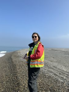 Someone stands holding a notebook and wearing a bright yellow vest. They are in a gravelly field with some tire tracks. To the left of the image, the ocean can be seen crashing into the gravelly beach. The sky is mostly blue with a hazy white are on the right of the image.