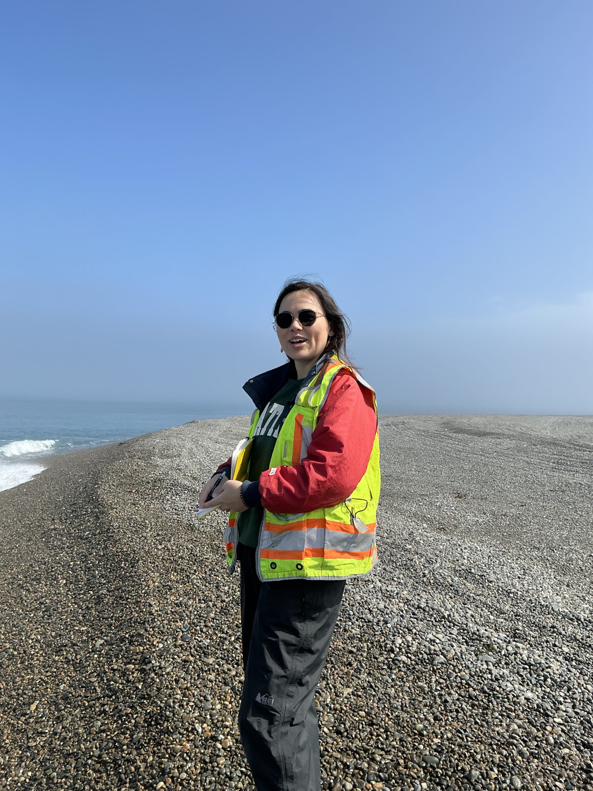 Someone stands holding a notebook and wearing a bright yellow vest. They are in a gravelly field with some tire tracks. To the left of the image, the ocean can be seen crashing into the gravelly beach. The sky is mostly blue with a hazy white are on the right of the image.