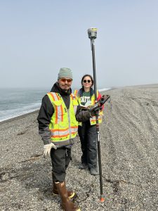 Two people stand next to each other. One is holding a Trimble instrument. They are standing on a gravelly beach. The water is lapping the shore on the left of the photo. The sky is grey and misty.