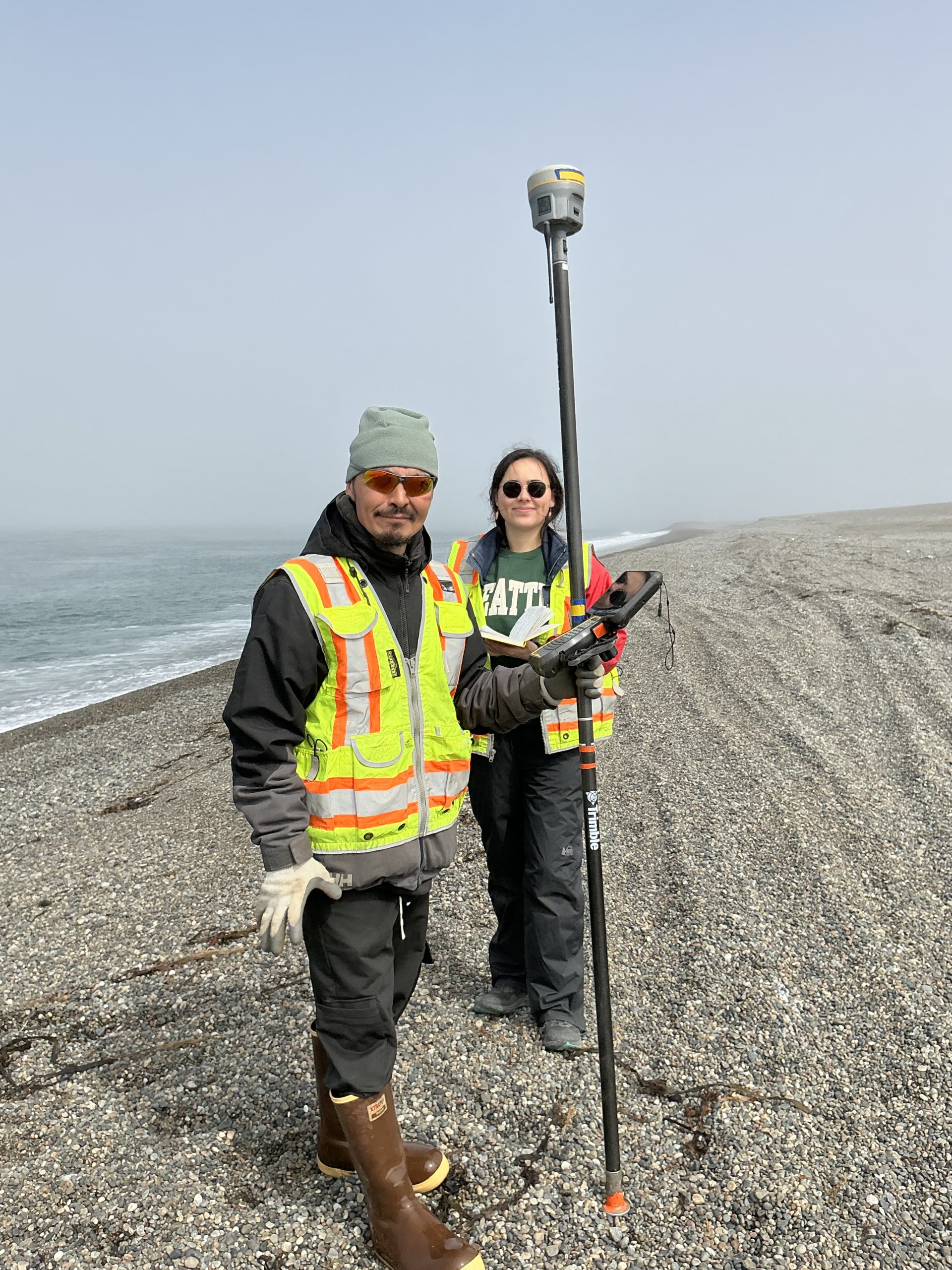 Two people stand next to each other. One is holding a Trimble instrument. They are standing on a gravelly beach. The water is lapping the shore on the left of the photo. The sky is grey and misty.