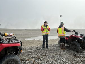 Two people standing on a gravelly field. One is holding a notebook and the other is holding a Trimble instrument. There are two ATVs on both sides of the photo. There is a pond behind them. The sky is light-grey.