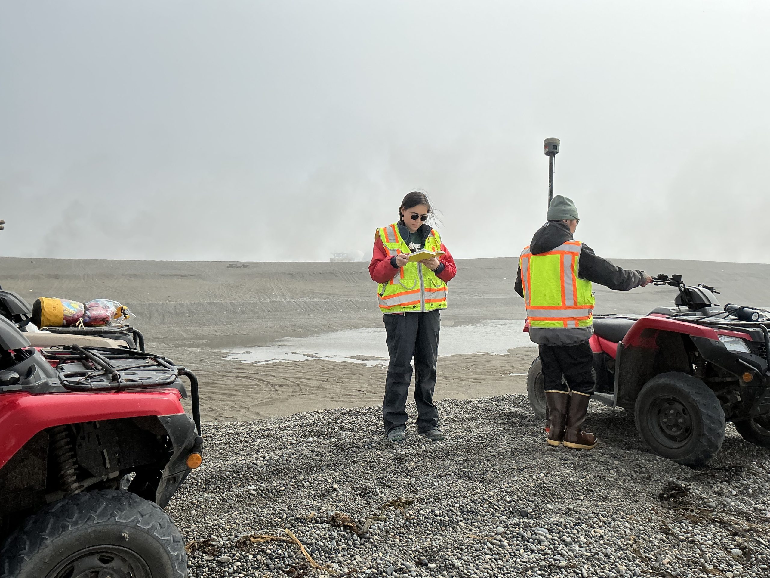 Two people standing on a gravelly field. One is holding a notebook and the other is holding a Trimble instrument. There are two ATVs on both sides of the photo. There is a pond behind them. The sky is light-grey.