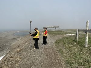 Two people stand on a gravelly hill facing the left of the frame. At the bottom of the hill, to the left of the frame, is a gravelly beach. Behind them, on the right of the frame, is a grassy field with a few gravelly roads. There is a worn wooden fence on the right of the frame. Behind the people is the skeleton of a canoe, flipped upside down, and propped up by wooden poles. The sky is misty grey.