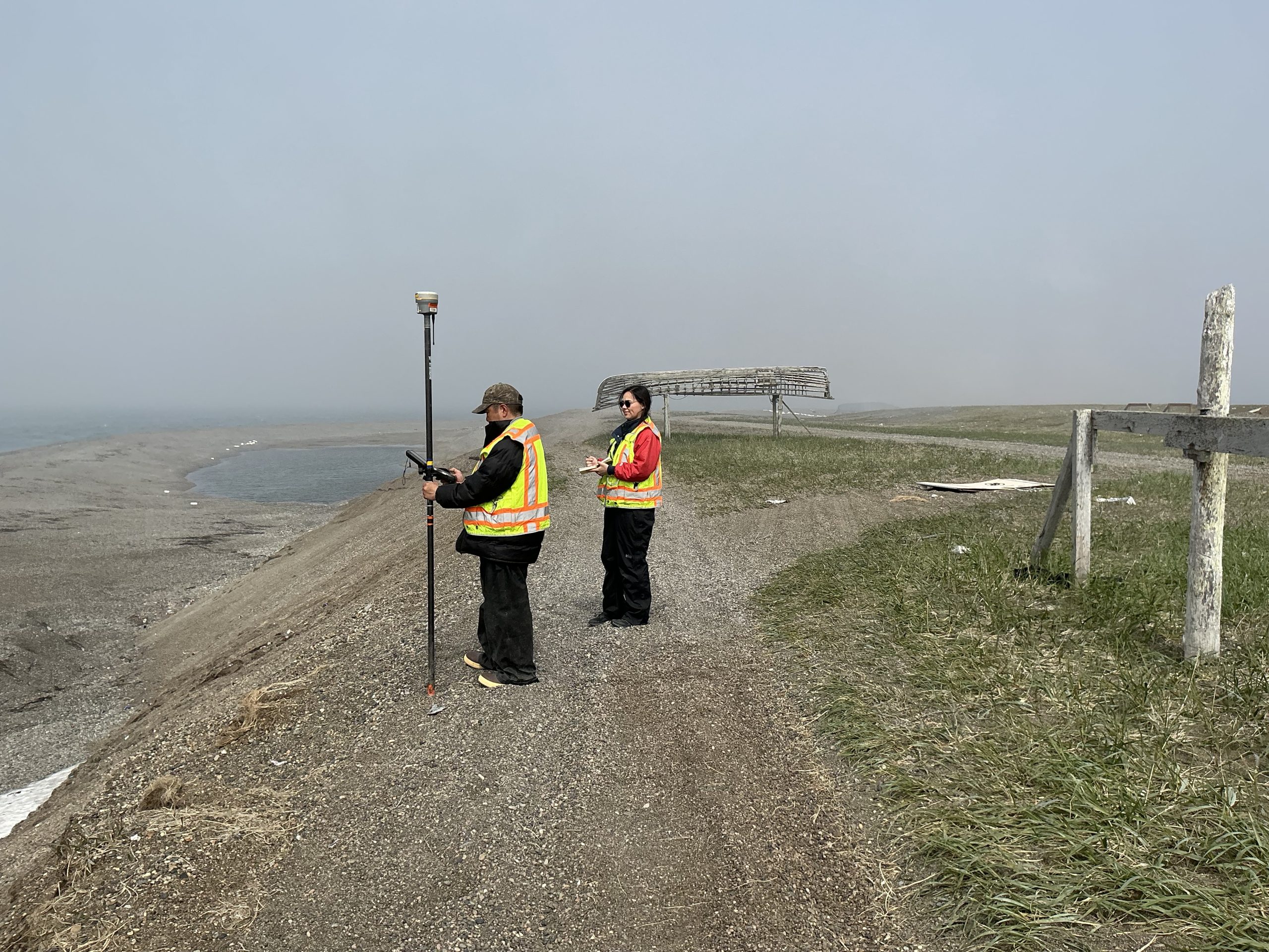 Two people stand on a gravelly hill facing the left of the frame. At the bottom of the hill, to the left of the frame, is a gravelly beach. Behind them, on the right of the frame, is a grassy field with a few gravelly roads. There is a worn wooden fence on the right of the frame. Behind the people is the skeleton of a canoe, flipped upside down, and propped up by wooden poles. The sky is misty grey.