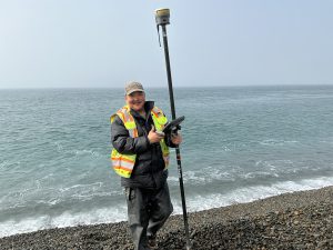 Someone stands near the beach's edge on a gravelly beach. They are holding a Trimble instrument. The water is calm behind them. The sky is a muted blue.
