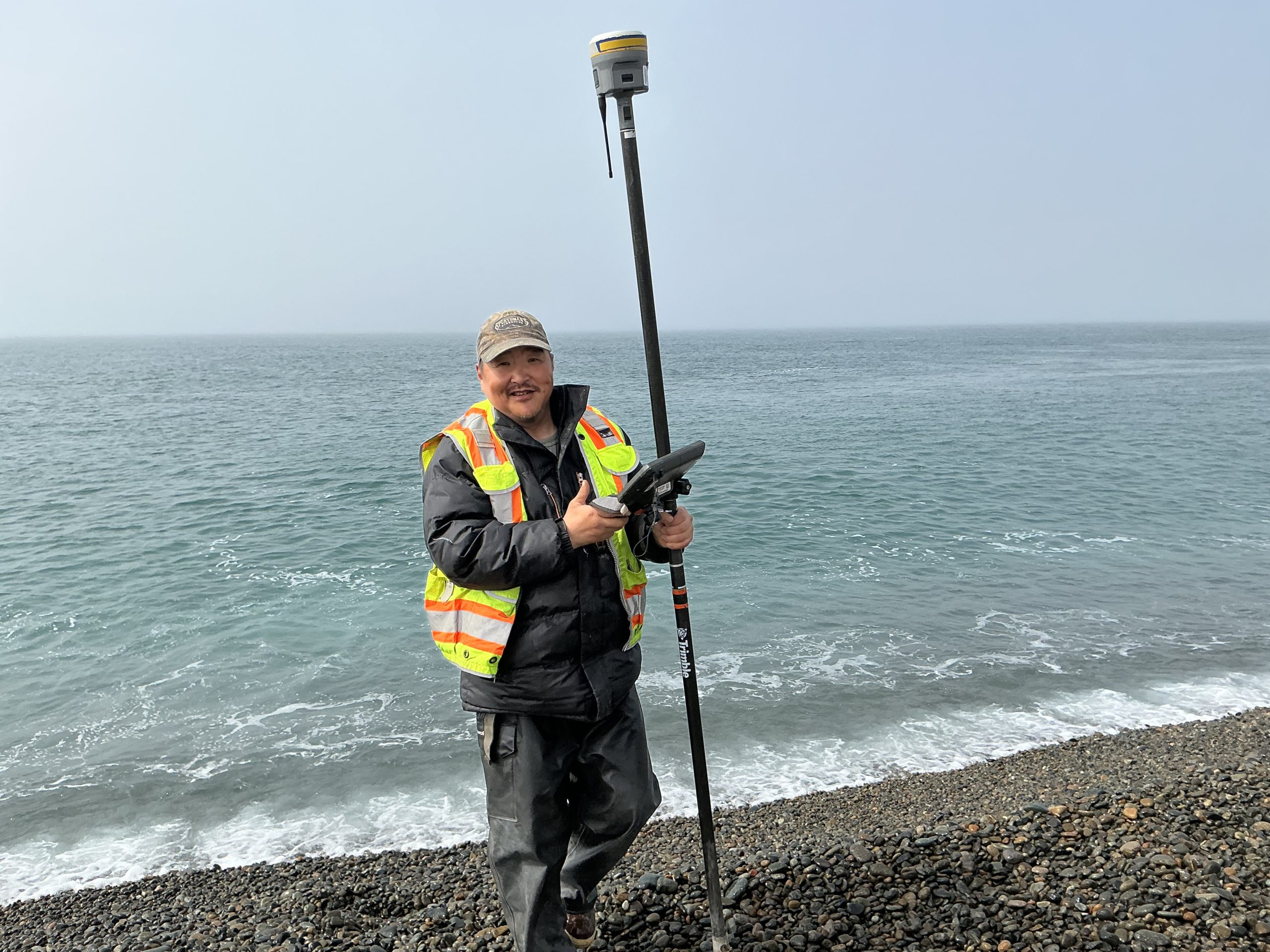 Someone stands near the beach's edge on a gravelly beach. They are holding a Trimble instrument. The water is calm behind them. The sky is a muted blue.