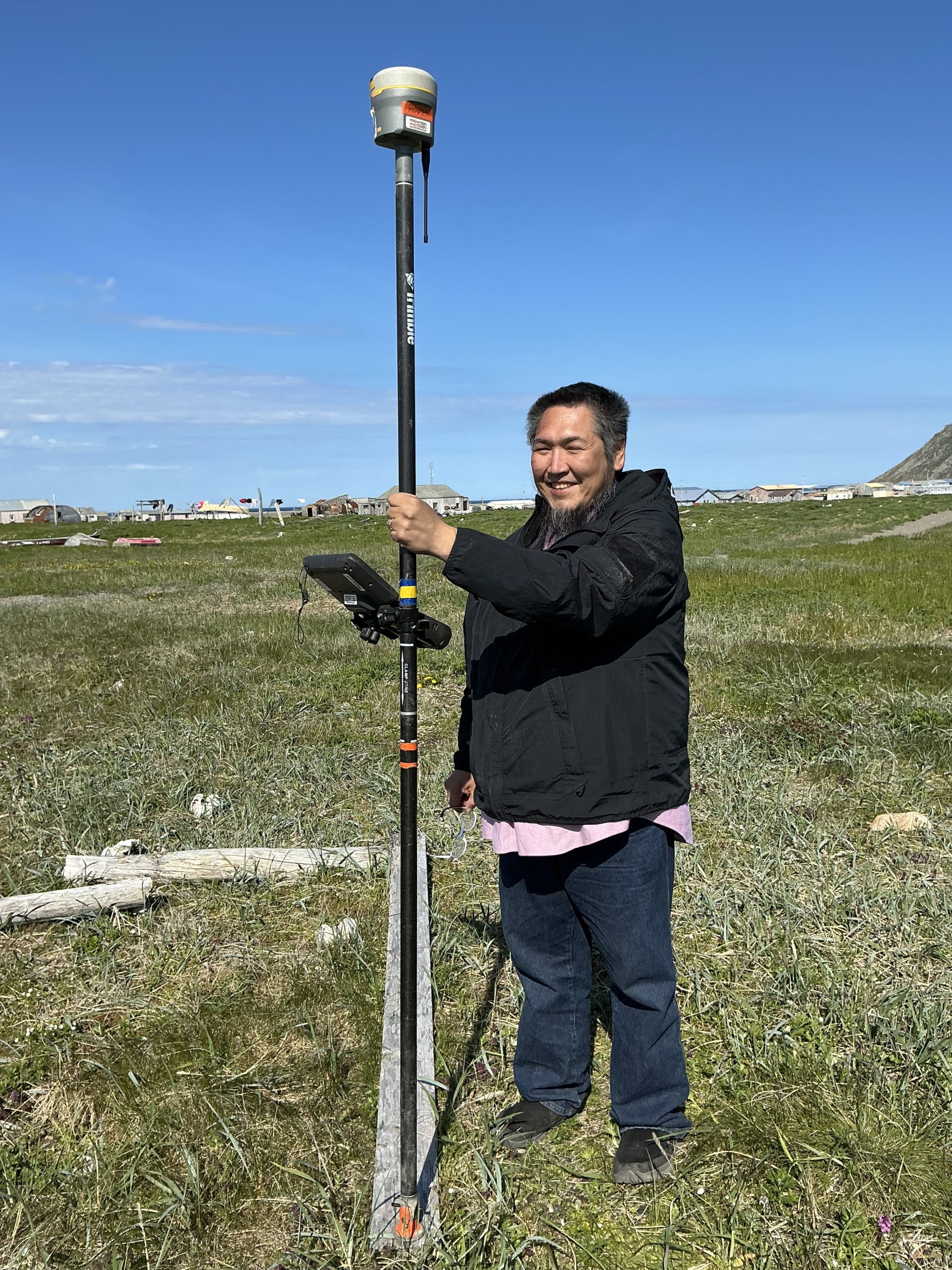 Someone stands in a grassy field holding a Trimble instrument. In the horizon are houses. The sky is blue with a couple of clouds.