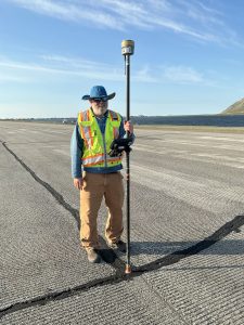 Chris stands in a paved road holding a Trimble instrument. In the background is dark blue water. The sky is blue with whispy clouds.