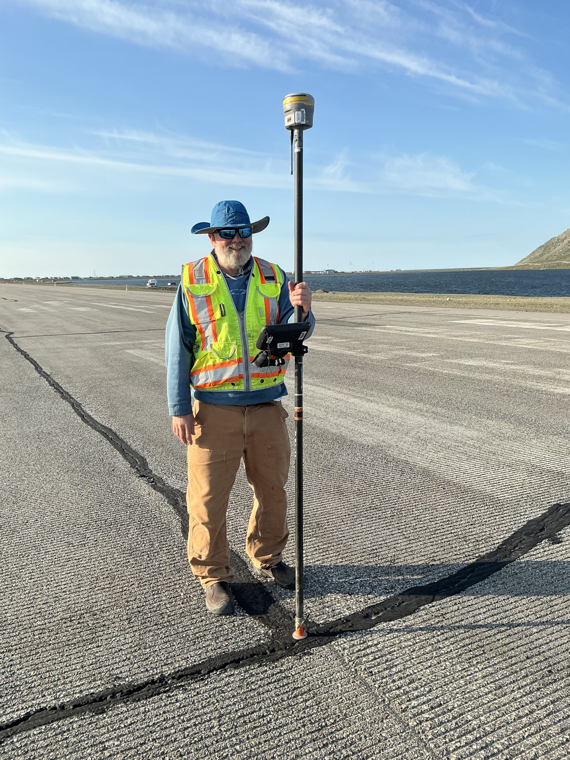 Chris stands in a paved road holding a Trimble instrument. In the background is dark blue water. The sky is blue with whispy clouds.