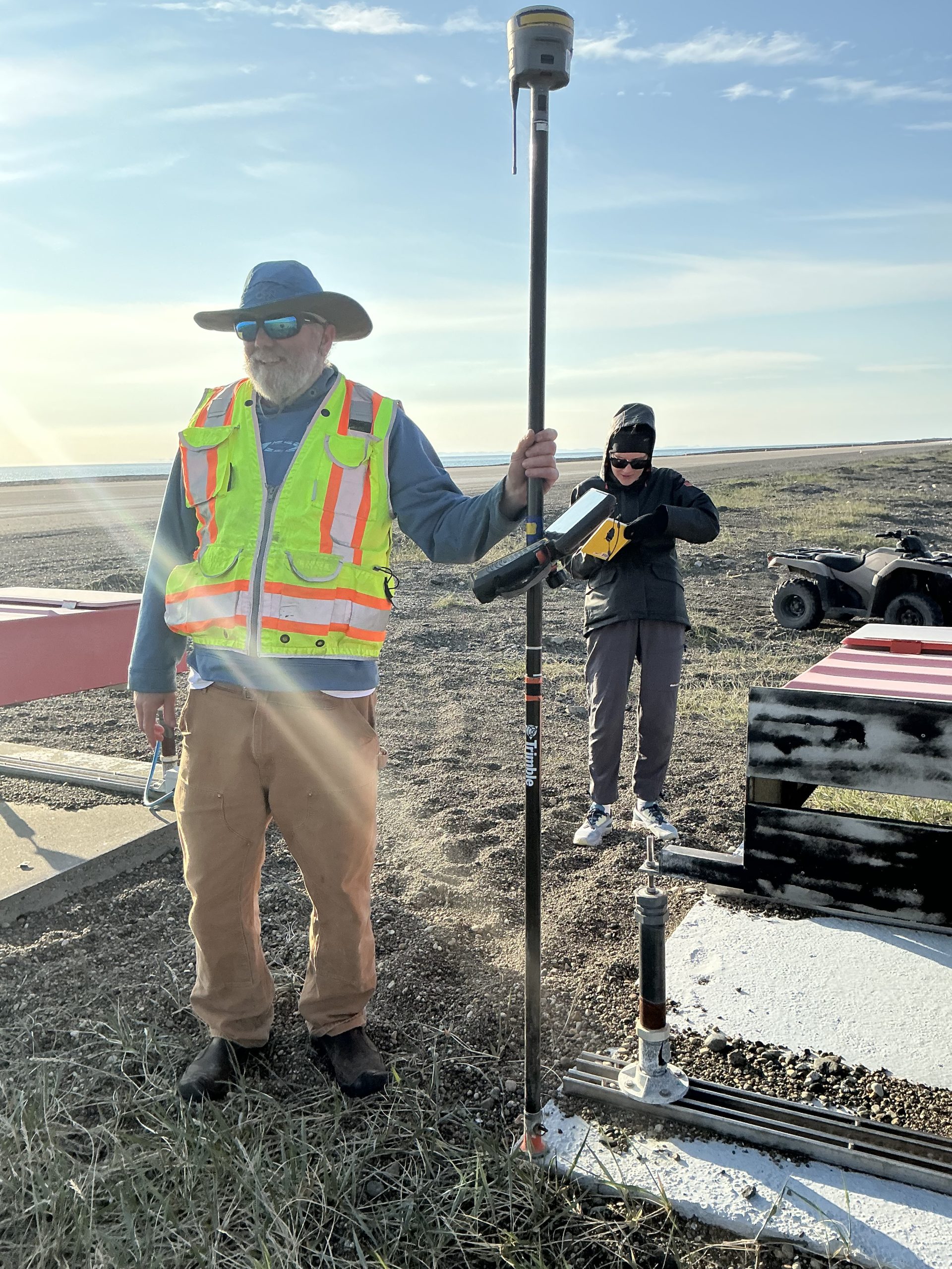 Chris stands in a gravelly patch holding a Trimble instrument. Behind him someone stands taking notes. There is an ATV further back. The ocean is barely visible in the horizon. The sky is blue and partially cloudy.