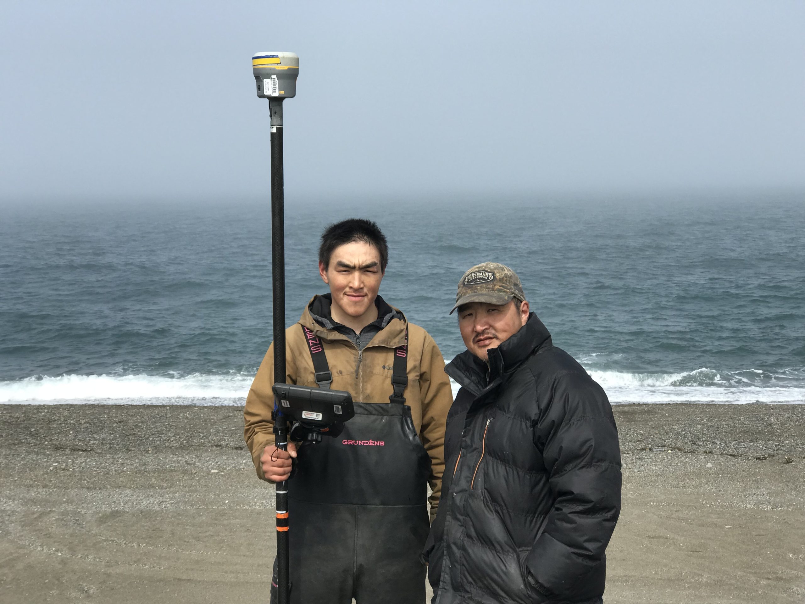 Two people stand next to each other. One is holding a Trimble instrument. They are standing on a gravelly beach. Behind them the ocean is lapping the shore. The water is a stormy blue. The horizon fazes into the misty sky.