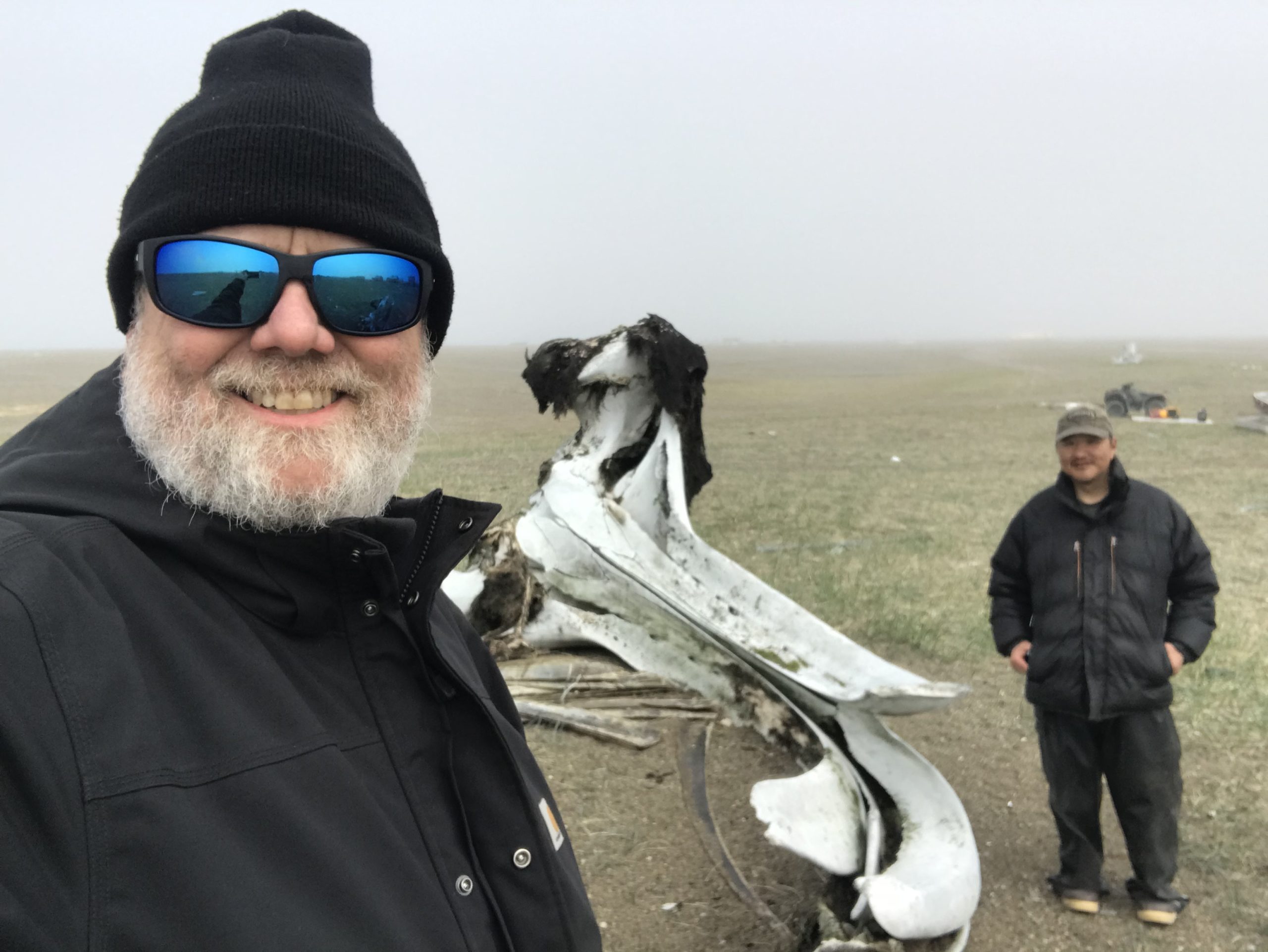 Two people stand in frame. One is holding the camera for a selfie, the other is further back, facing the camera and they are both smiling. Between them is a whale skull. They are standing in a partially dry grassy field. The sky is misty and grey.
