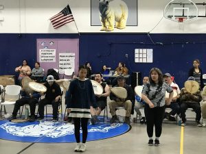 A group of people stand and sitting in a gymnasium. Some are holding drums and singing. Two are standing in front of the people with drums and appear to be dancing.
