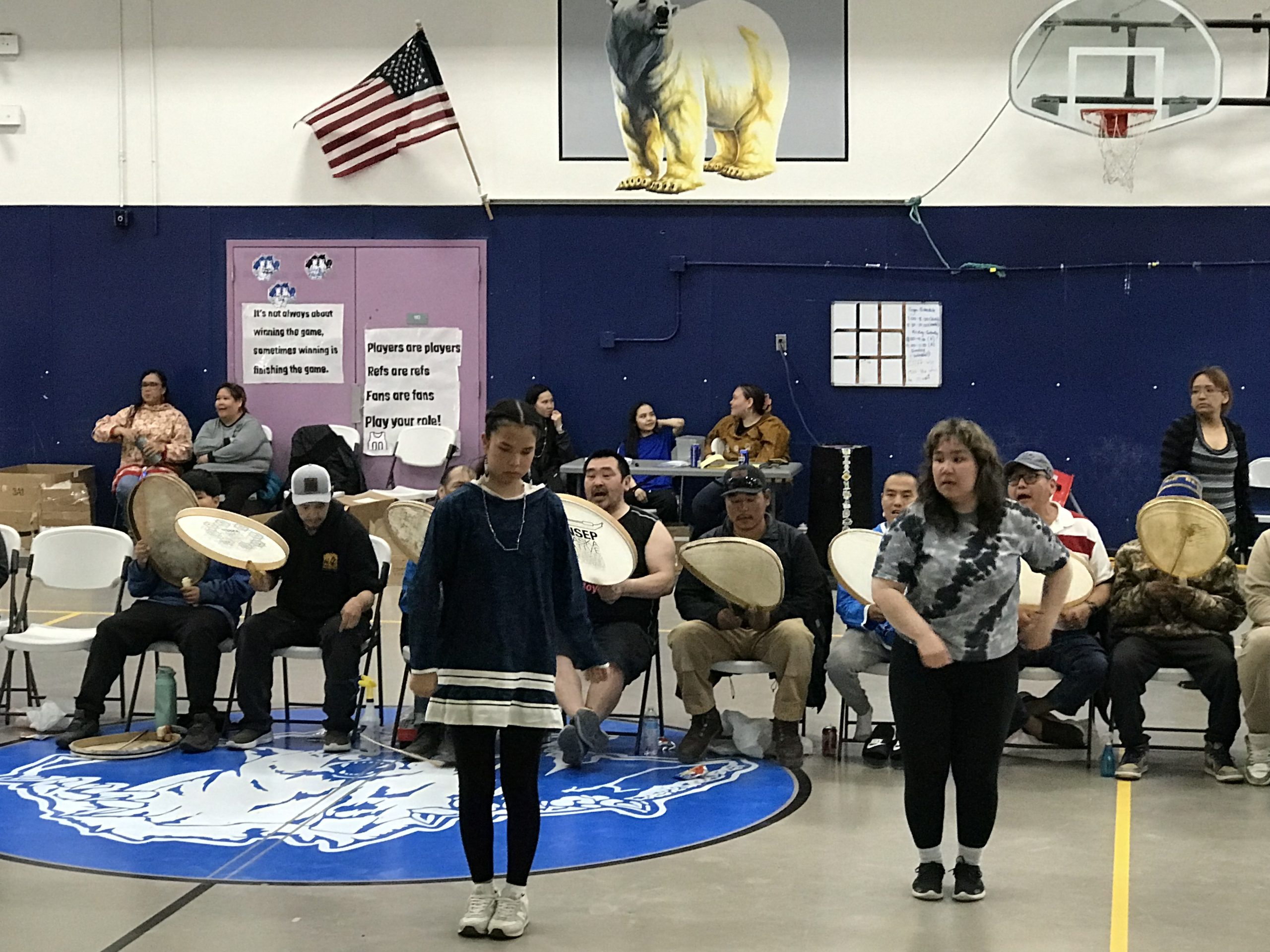 A group of people stand and sitting in a gymnasium. Some are holding drums and singing. Two are standing in front of the people with drums and appear to be dancing.