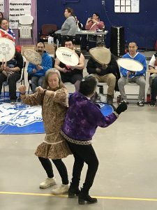 A group of people standing and sitting in a gymnasium. A line of people are holding drums and singing. Two people are hip-bumping and wearing some traditional clothing.