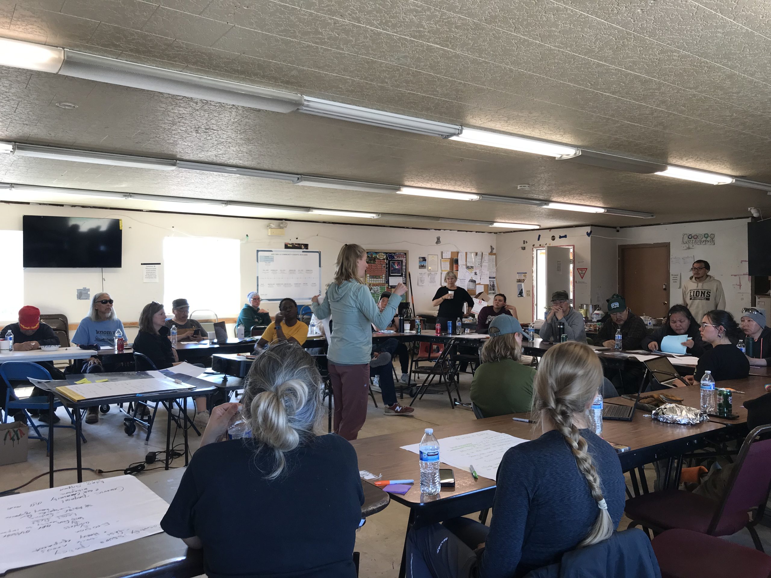 Someone stands in the middle of a class room. Tables are arranged in a U-shape around the person. People sit at the tables facing the person in the center, listening to what they have to say.