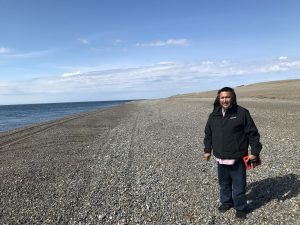 Someone stands in a gravelly beach. The ocean is calm and to the left of the frame. The ground slopes uphill to the right. The sky is blue and partially cloudy.