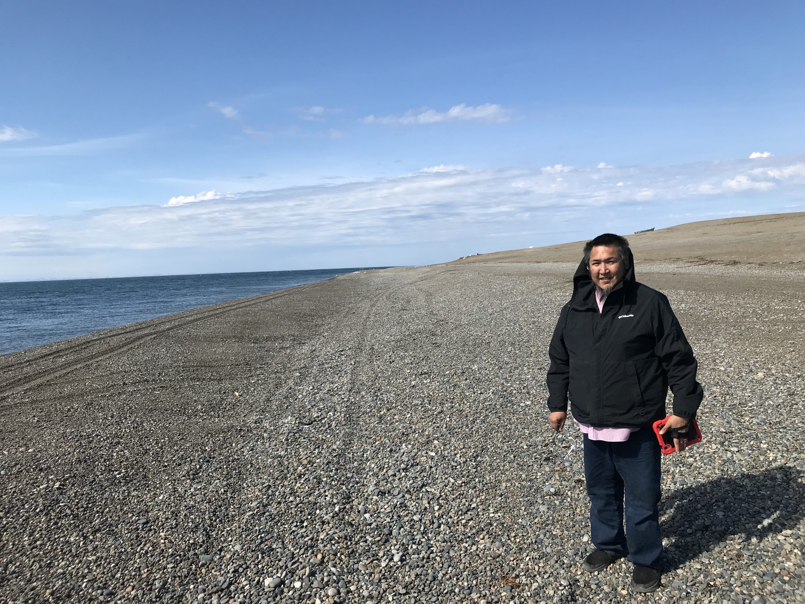 Someone stands in a gravelly beach. The ocean is calm and to the left of the frame. The ground slopes uphill to the right. The sky is blue and partially cloudy.