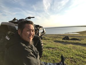 Someone is sitting next to an ATV smiling towards the camera. They are sitting on the top of a sloped hill looking down at calm waters. The sky is partially cloudy.