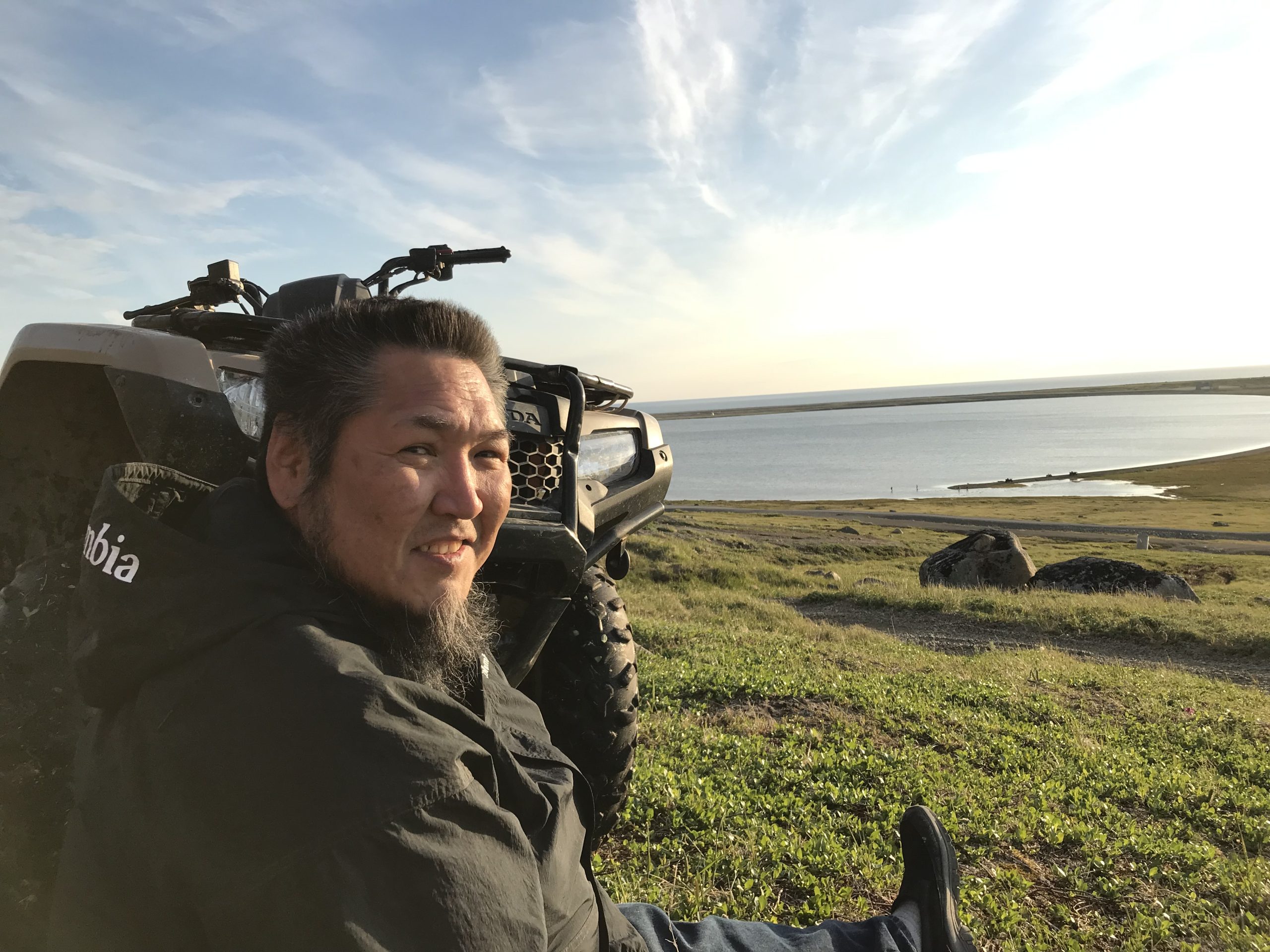 Someone is sitting next to an ATV smiling towards the camera. They are sitting on the top of a sloped hill looking down at calm waters. The sky is partially cloudy.