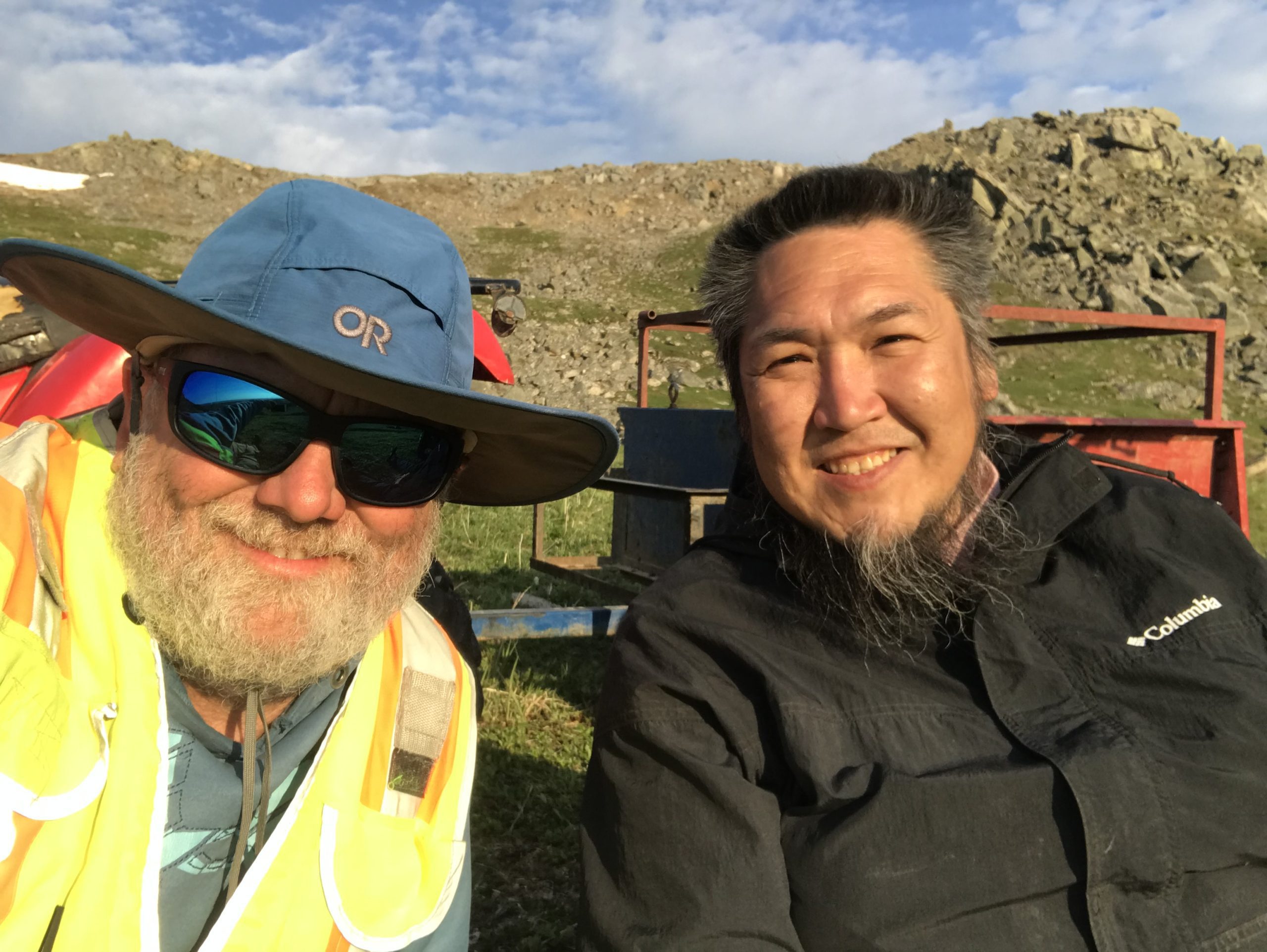 Two people sitting together for a selfie. There is a trailer behind them. They are sitting in a grassy field. Further back is a hill with some rocks piled on. The sky is blue and partially cloudy.