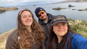 Three people standing close together for a group selfie, there is a small hill behind them and smooth waters beyond the hill.