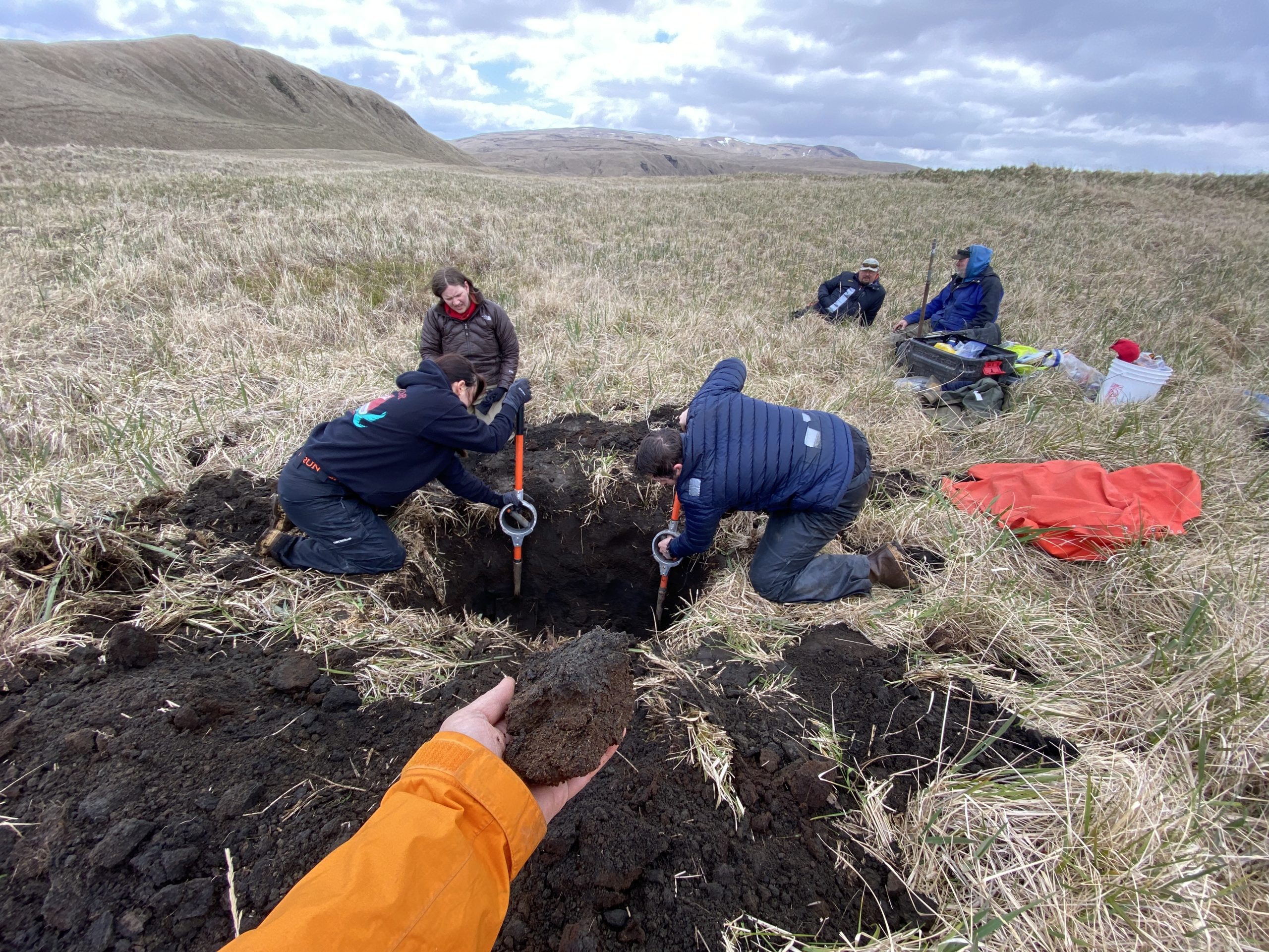 A grassy field with hills in the background. A hand extends from the bottom of the photo holding a fist-sized clump of dirt, two other people are digging a deep hole, one person sits on the side of the hole looking at the two digging, and two people a little further back sit and chat with each other.