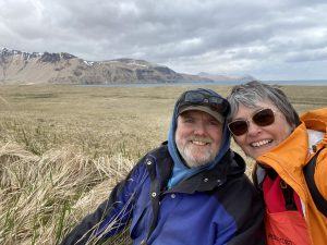Piama and Chris smiling for a selfie. Behind them is a dried grassy field. The background shows mountains and a cloudy, overcast sky.