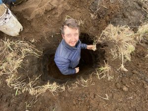 Reyce stands in a freshly dug hole in the ground looking up to the camera, he is holding a clump of dried grass in his left hand.