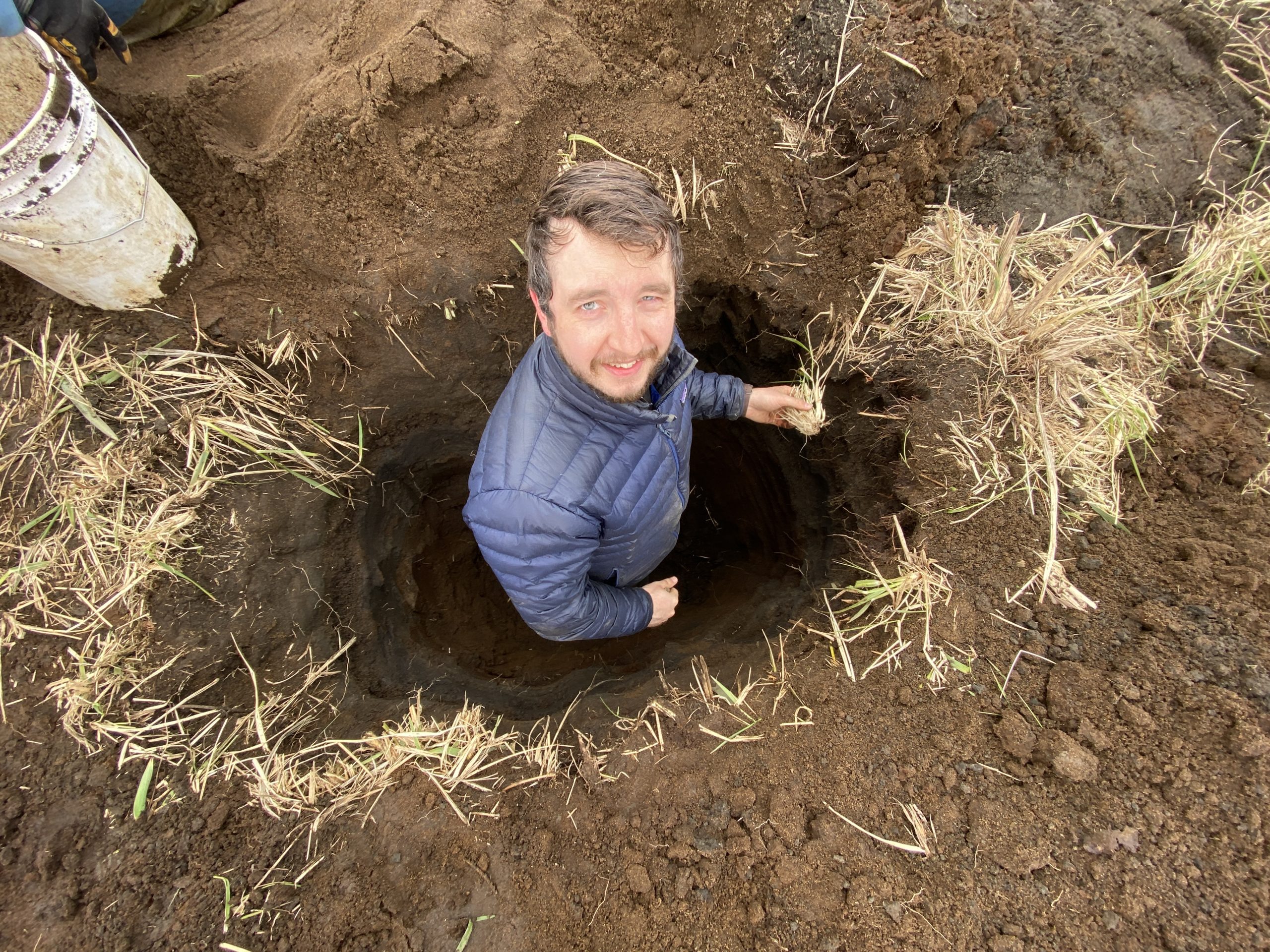 Reyce stands in a freshly dug hole in the ground looking up to the camera, he is holding a clump of dried grass in his left hand.