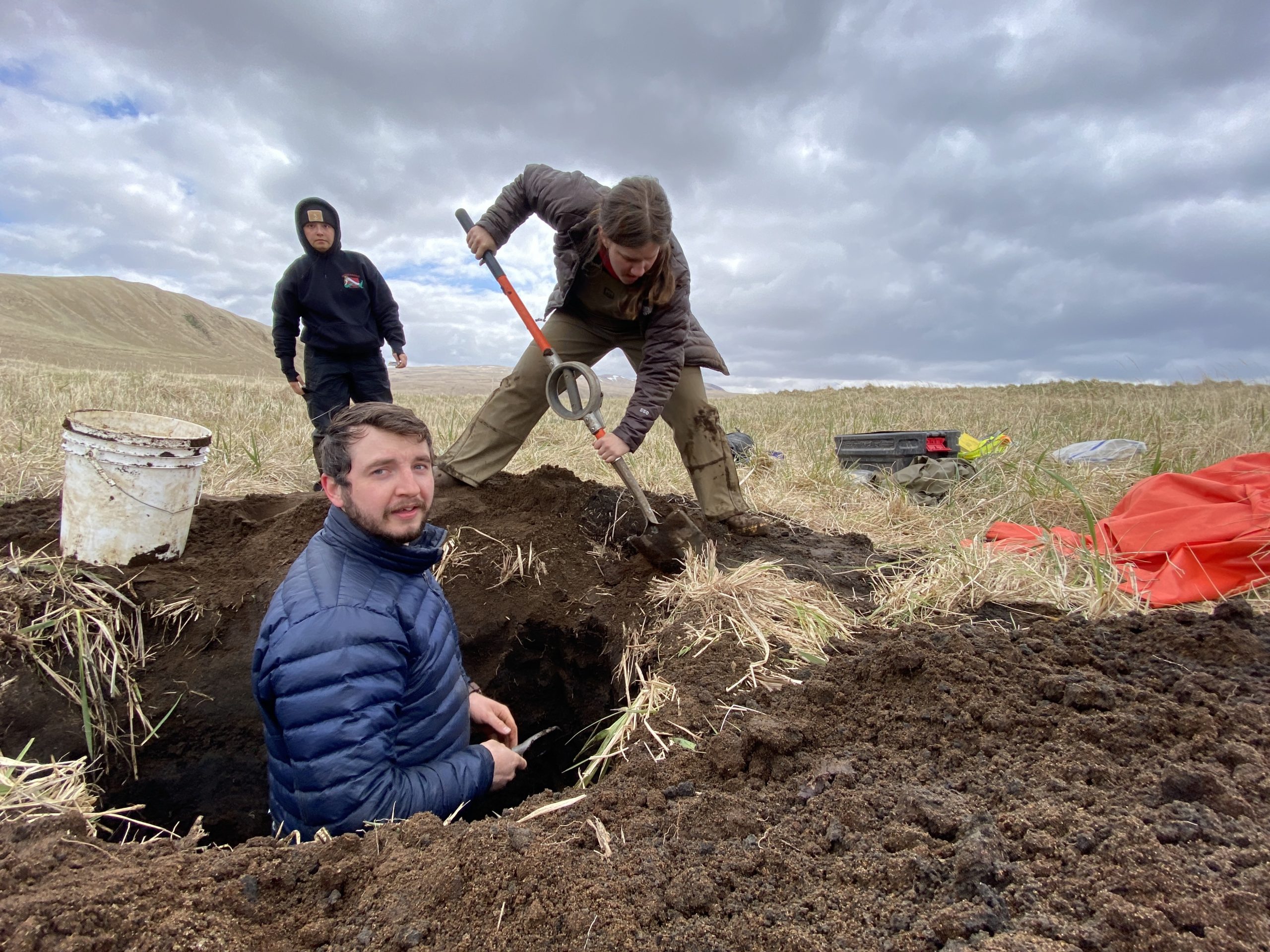 One person is standing in a freshly dug hole, another person is digging around the hole, and a third person walks towards the hole. The sky is overcast and the background is hilly with dried grass.