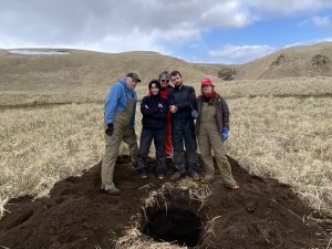 Five ACGL members stand behind a freshly dug hole trying to look tough (they're not). The background is dried grass and hilly with a partially cloudy sky