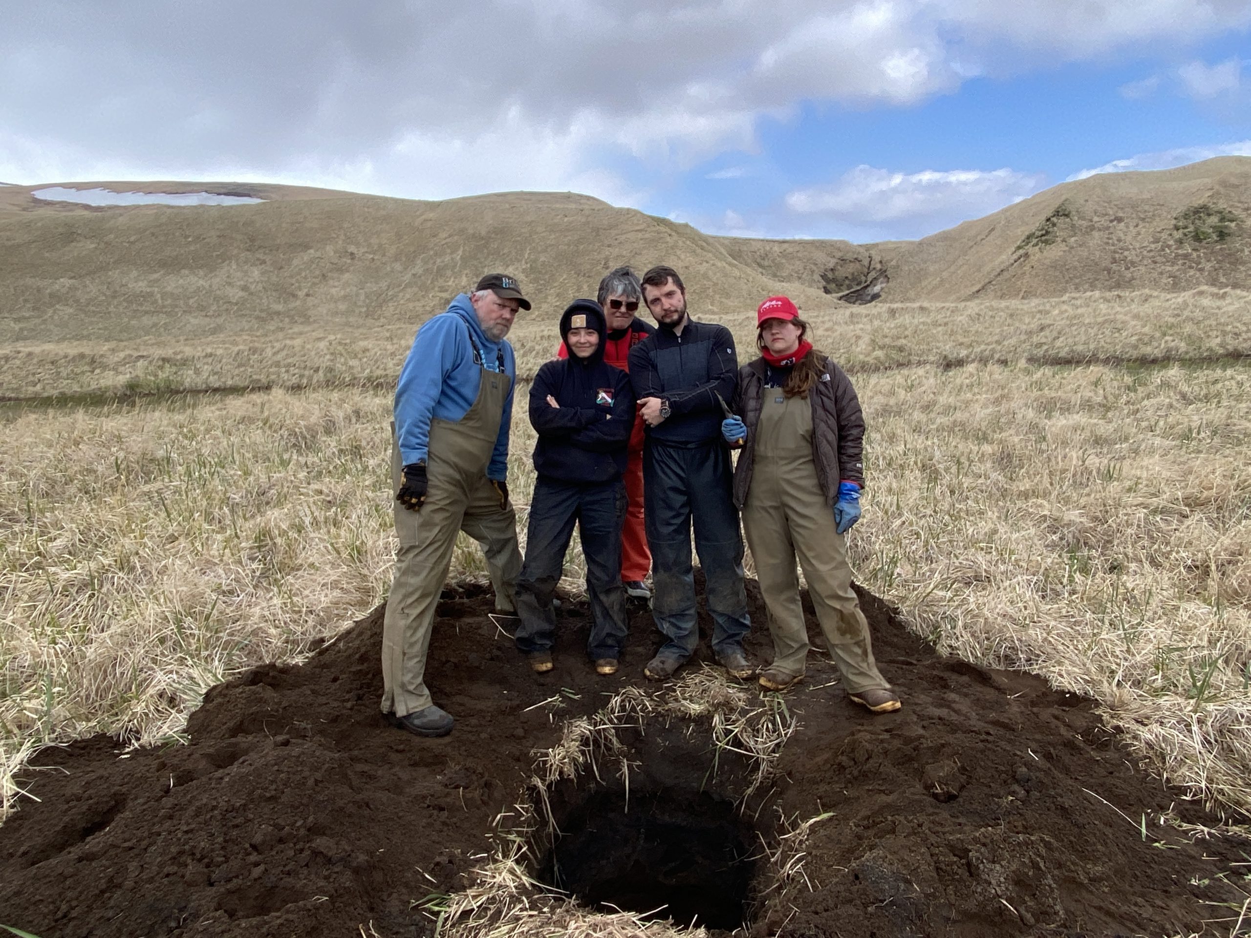 Five ACGL members stand behind a freshly dug hole trying to look tough (they're not). The background is dried grass and hilly with a partially cloudy sky