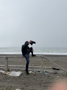 Someone leans on a wooden fence for support. They are holding one of their shoes upside down and water is dripping out of the shoe. They stand on a gravelly beach. Behind them the water is lapping the shore. The sky is misty and grey.