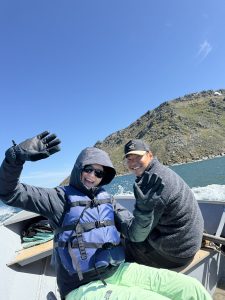 Two people sitting in a boat. Water is splashing around the outside of the boat. The water appears calm and blue. There is a rocky, grassy hill in the background. The sky is blue.