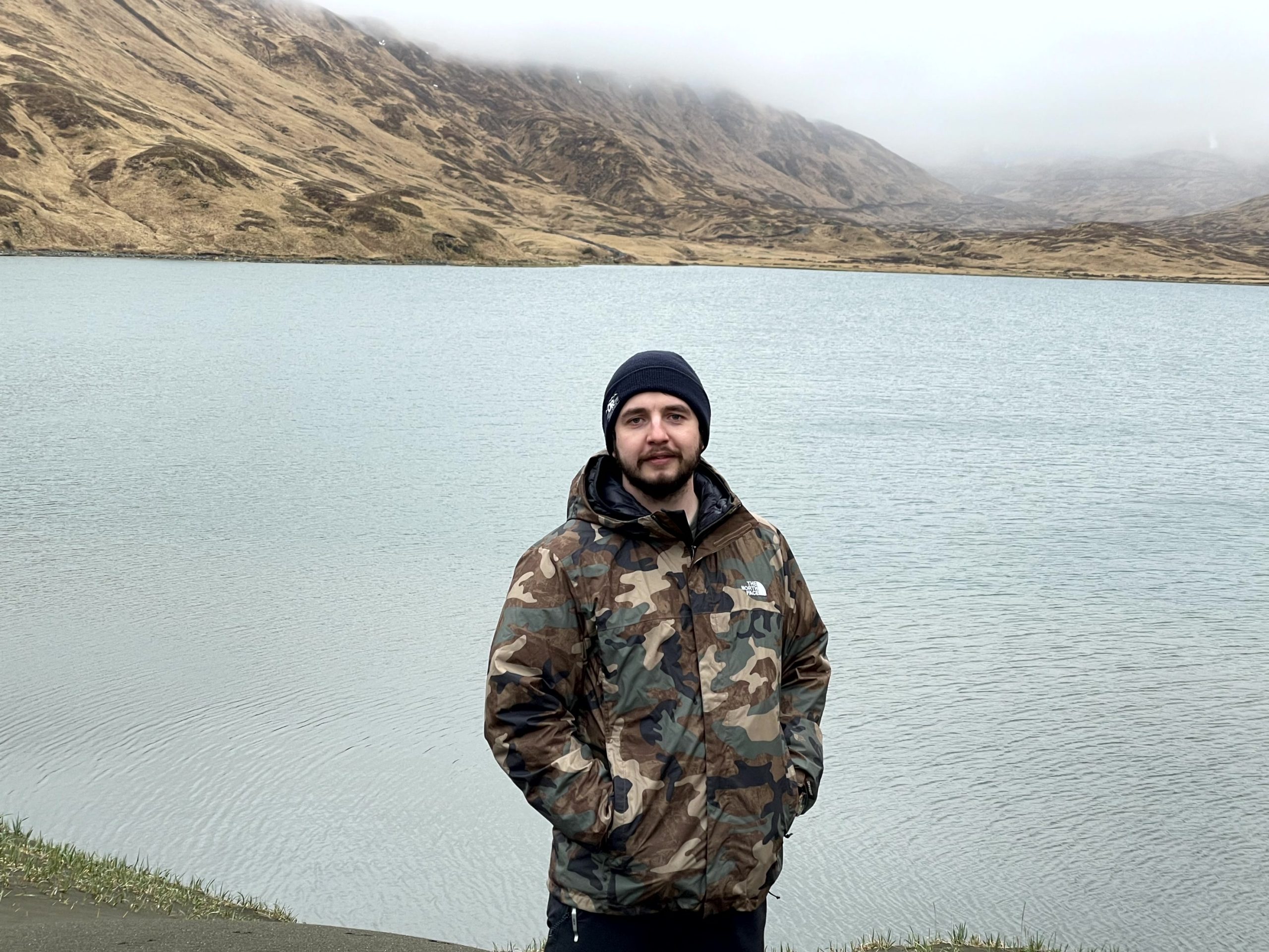 Reyce stands in front of a calm lake, the hills in the background are capped with misty, low lying clouds.