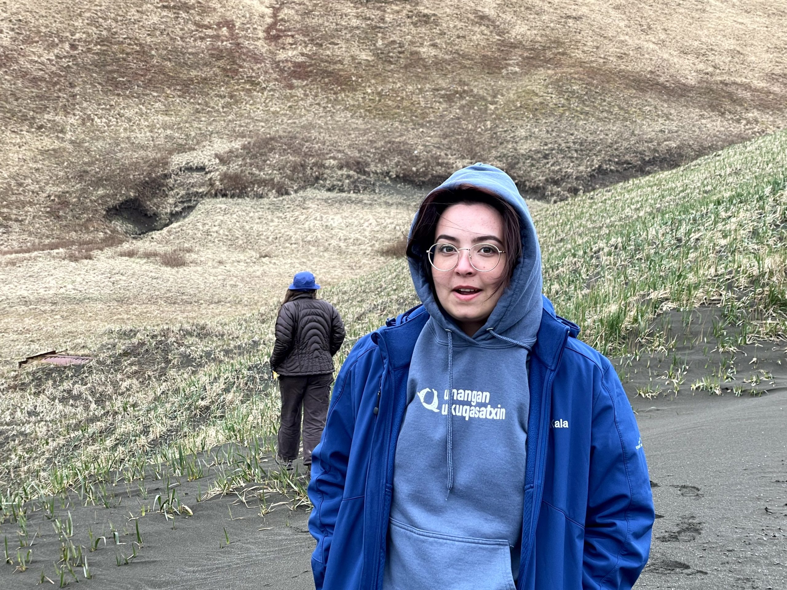 One person stands closer, looking at the camera, another person is in the background walking away from the camera. The environment is hilly grass.