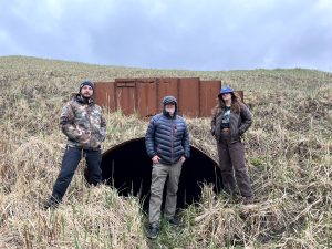 Three people stand in front of a large drainage tunnel. The background is a grassy hill and the overcast sky is light purple and blue
