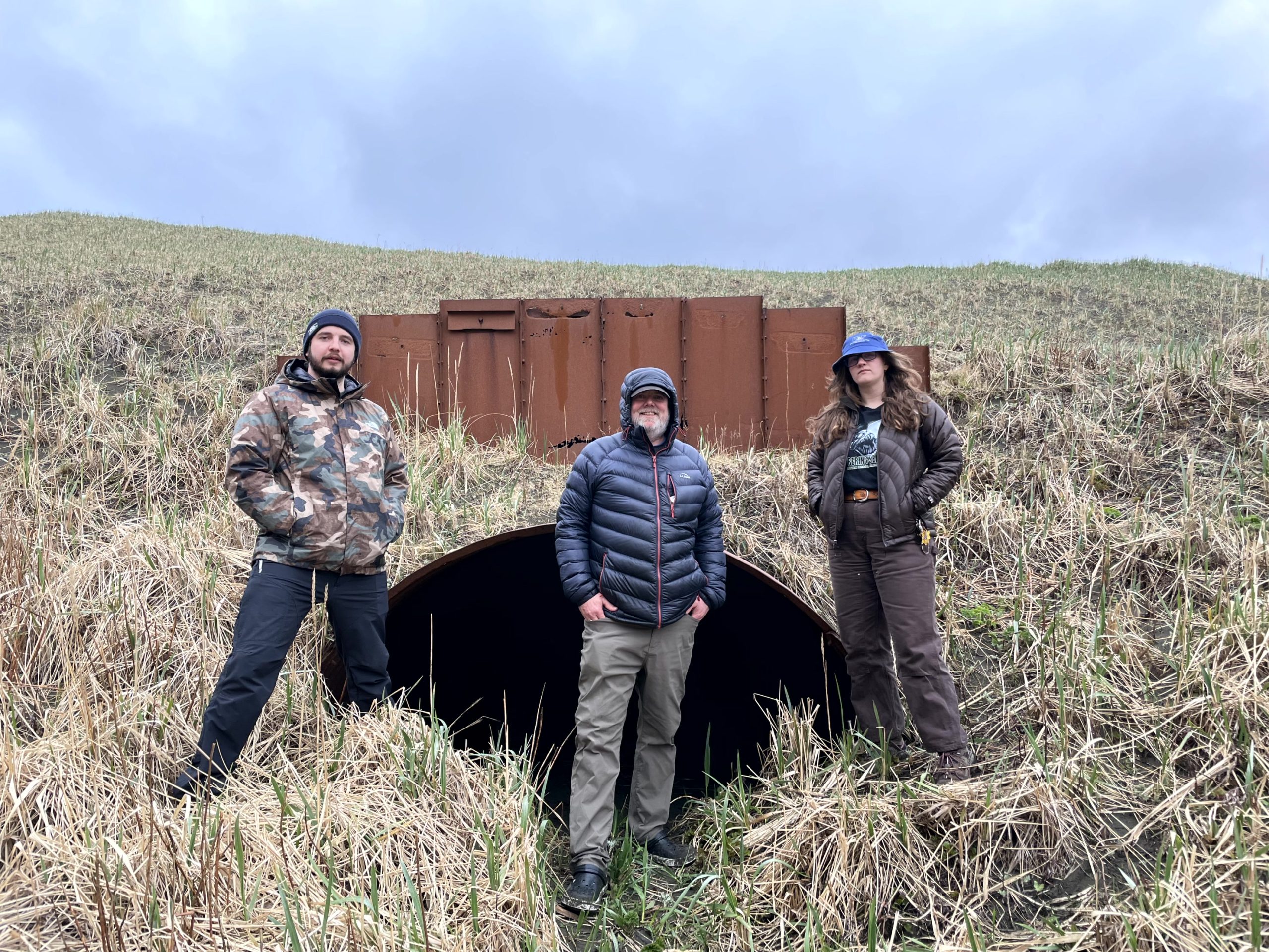 Three people stand in front of a large drainage tunnel. The background is a grassy hill and the overcast sky is light purple and blue
