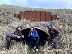 Three people squatting in front of a large storm drain. The background is a grassy hill with light bluish-purple clouds