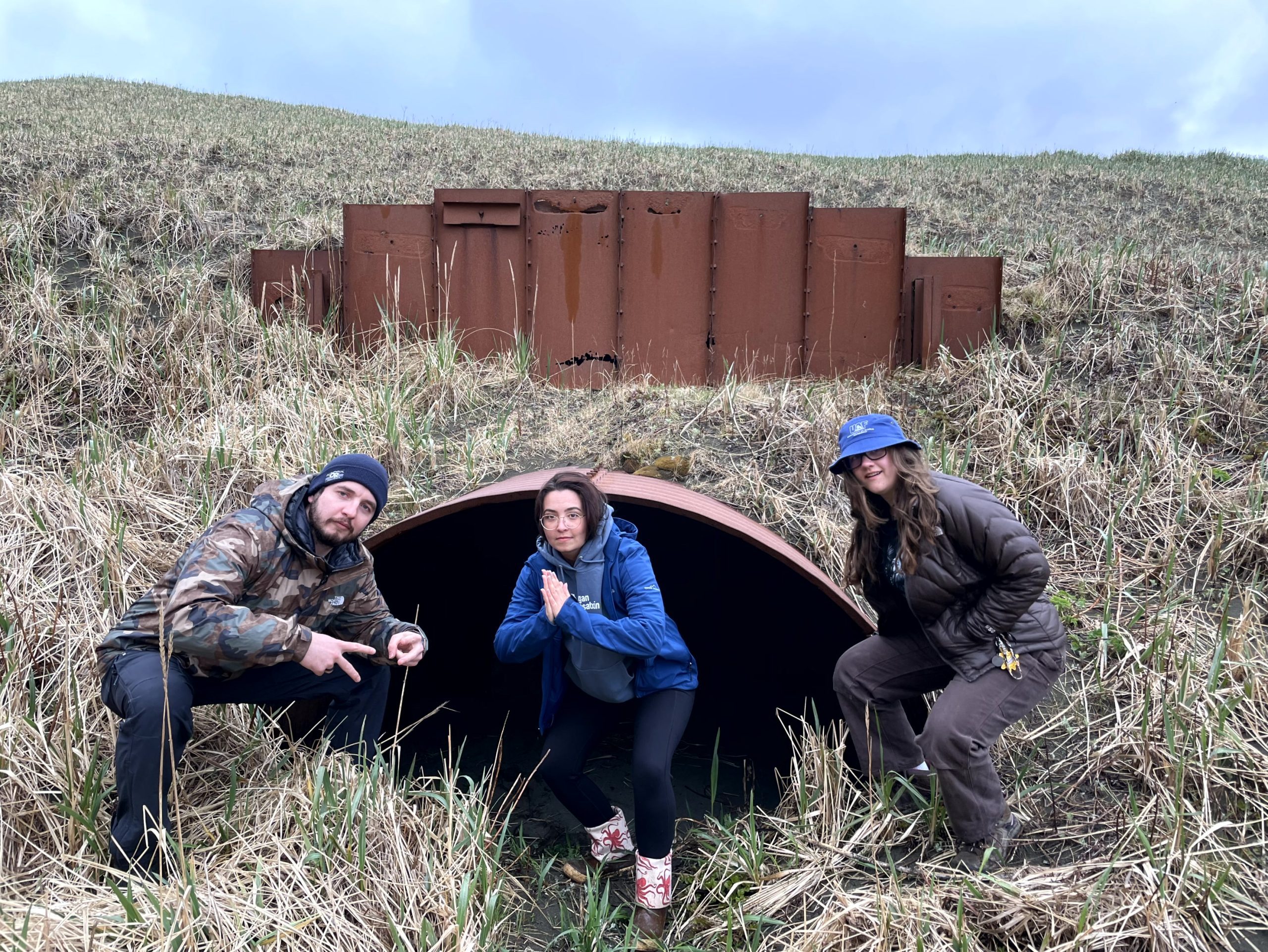 Three people squatting in front of a large storm drain. The background is a grassy hill with light bluish-purple clouds
