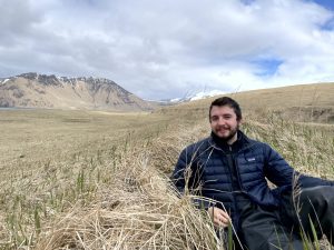 Reyce is half-sitting half-lying down on a grassy hill with a mountain in the background and a partially cloudy sky