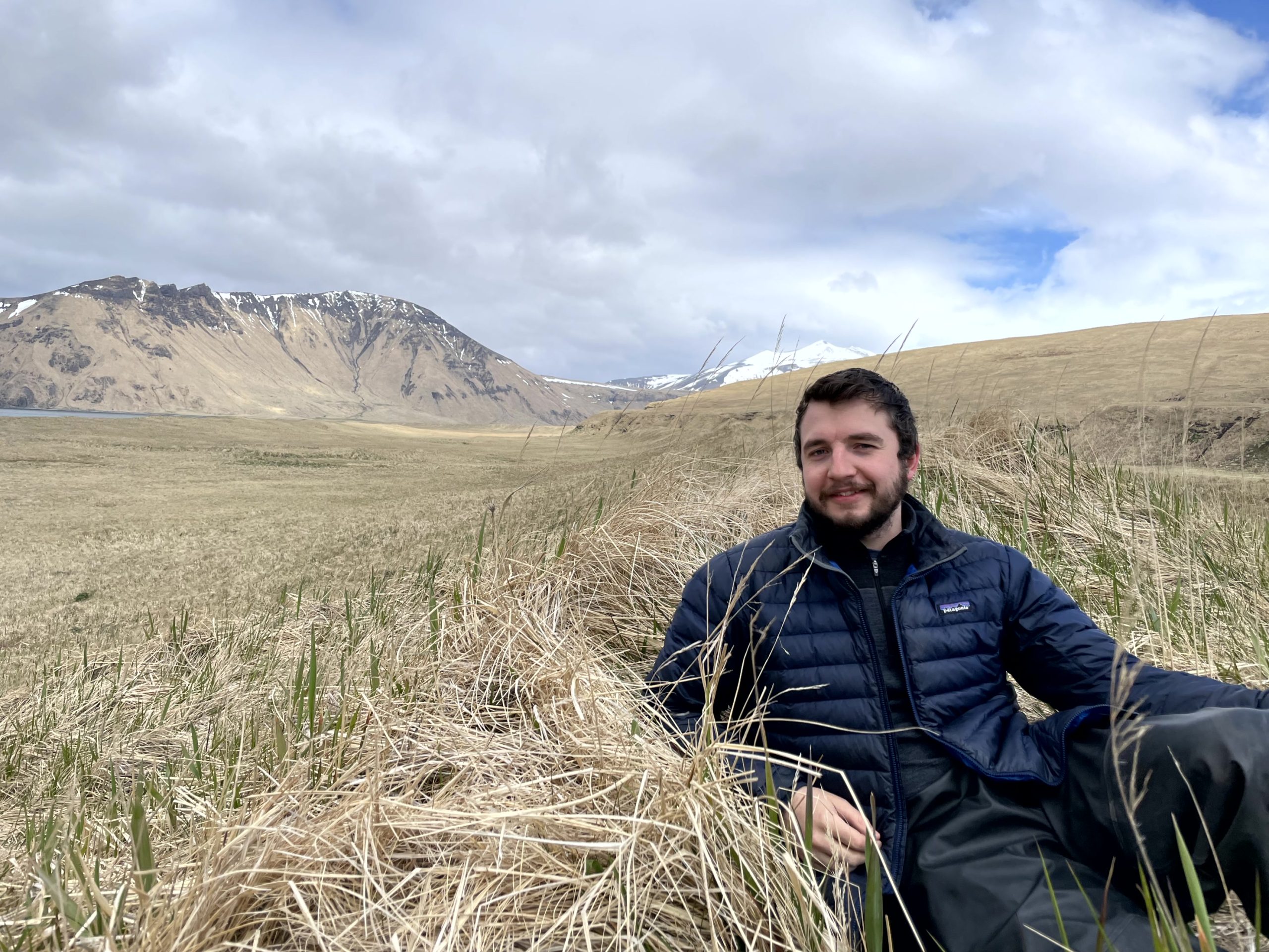Reyce is half-sitting half-lying down on a grassy hill with a mountain in the background and a partially cloudy sky
