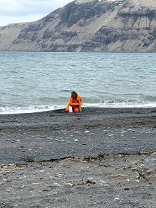 Piama in a bright orange outfit kneels on a rocky beach with a small, white container in front of them. The ocean is breaking right behind her and the water is calm. The bottom part of a mountain can be seen in the background. The sky is overcast.