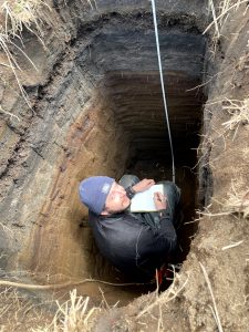 Reyce is crouched in a deep hole looking up to the camera and writing in a field notebook