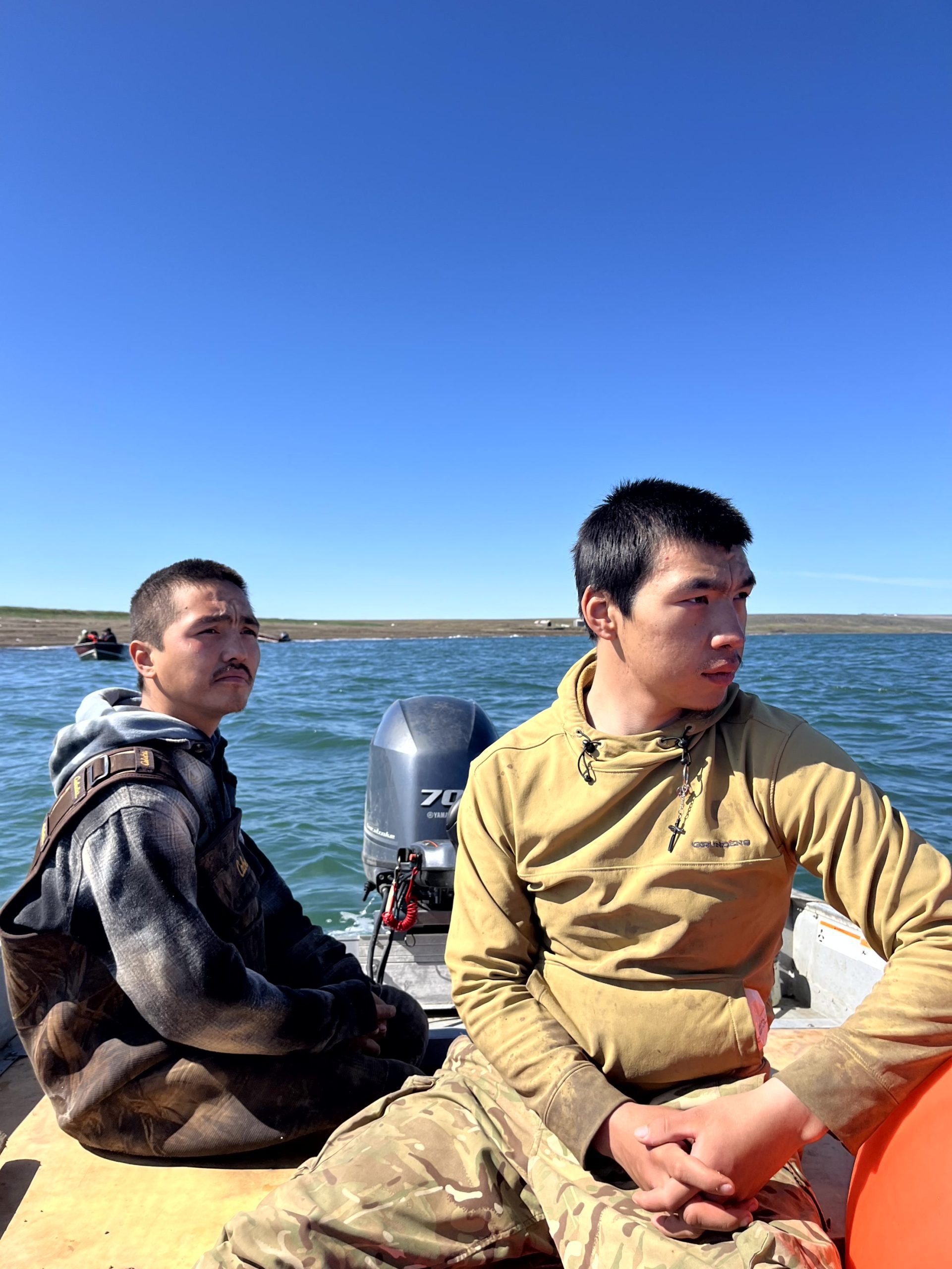 Two people sit in a boat looking off to the right. The water around them is calm and blue. The land can be seen along the horizon. The sky is clear and a deep blue.